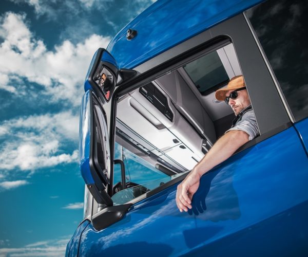 Heavy Load Cargo Delivery. Caucasian Truck Driver Inside Semi Tractor Cabin and the Blue Sky. Transportation Industry.