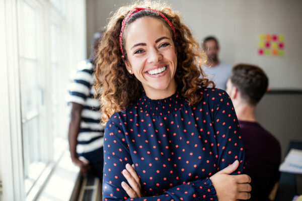 Young female designer smiling while standing with her arms crossed after an office meeting with colleagues standing in the background