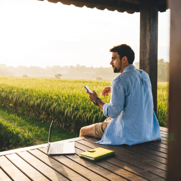 Side view of concentrated young male freelancer in shirt sitting near laptop on wooden podium in picturesque countryside and browsing smartphone during sunset