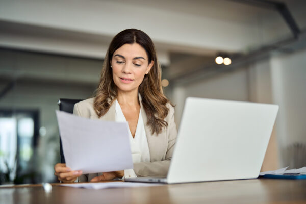 Busy 40 years old business woman working in office checking documents. Mid aged businesswoman accounting manager executive or lawyer using laptop reading paper file financial report, tax invoice.