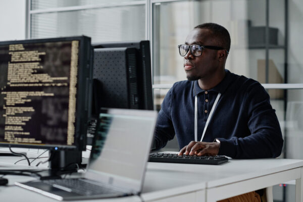 Developer working with new program African American young developer in eyeglasses concentrating on his online work on computer sitting at workplace