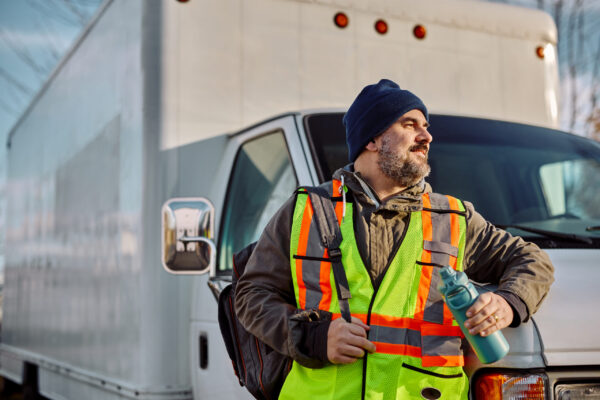 Happy truck driver with backpack and water bottle in front of his vehicle on parking lot.