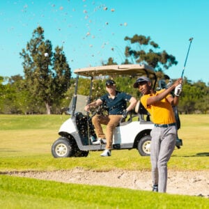 Caucasian young man sitting in cart looking at african american friend playing golf against sky Caucasian young man sitting in cart looking at african american friend playing golf against sky